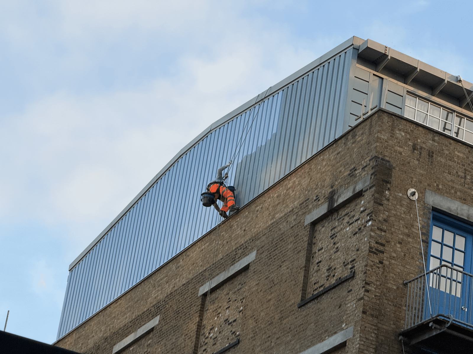 A painter working high on a building's blue cladding on ropes