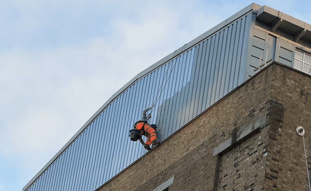 A worker on ropes along metal cladding high on a brick building